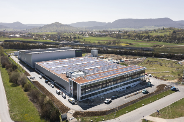 Luftbild einer großen Halle mit Solarmodulen auf dem Dach, umgeben von Fahrzeugen, Bäumen, Gras, Masten und Hügeln unter einem klaren blauen Himmel, identifiziert als Deutschlands erste solarbetriebene Fabrik.
