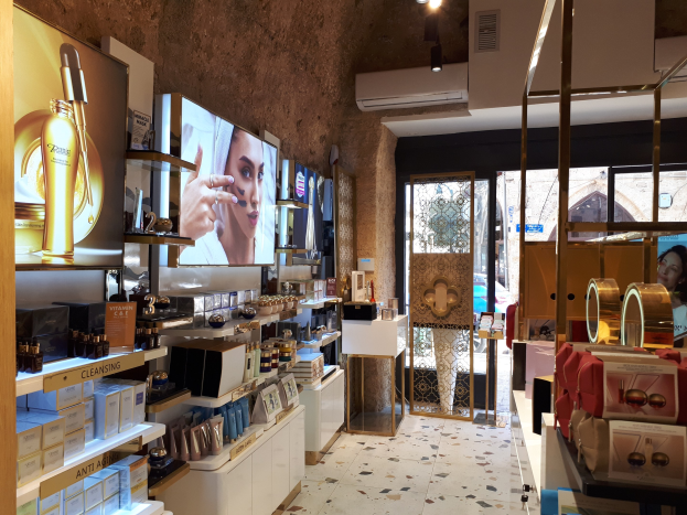 Ein Kosmetikgeschäftsinneres mit Regalen, die verschiedene Schönheitsprodukte zeigen, an der Wand montierte Bildschirme, Deckenleuchten, eine Klimaanlage und Glastüren, die eine Person, ein Auto und ein Gebäude zeigen.