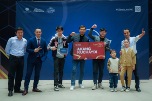 Eine Gruppe von Menschen auf einer Bühne, zwei halten eine Tafel mit dem Text "Akang Kuchaydi - Zakovat Quiz", mit einem Banner, Scheinwerfern und anderen Gegenständen im Hintergrund.