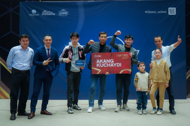 Eine Gruppe von Menschen auf einer Bühne, zwei halten eine Tafel mit dem Text "Akang Kuchaydi - Zakovat Quiz", mit einem Banner, Scheinwerfern und anderen Gegenständen im Hintergrund.