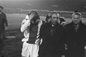 Schwarzes und weißes Bild einer Gruppe von Männern, die auf einem Fußballfeld stehen und nach einem Spiel besorgt aussehen, mit einem Stadion im Hintergrund.