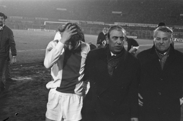 Schwarzes und weißes Bild einer Gruppe von Männern, die auf einem Fußballfeld stehen und nach einem Spiel besorgt aussehen, mit einem Stadion im Hintergrund.