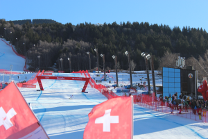 Eine Gruppe von Menschen steht auf einer schneebedeckten Hänge mit roten Fahnen im Vordergrund, Laternen, Tälern und einem klaren blauen Himmel im Hintergrund und einem großen Bildschirm auf der rechten Seite.