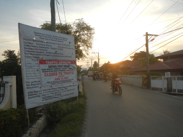 Eine Gruppe von Motorradfahrern auf einer Straße mit einem Schild "Verkehrshinweis Gesperrt für den Verkehr", gesäumt von einer blühenden Wand und Bäumen unter einem klaren blauen Himmel.