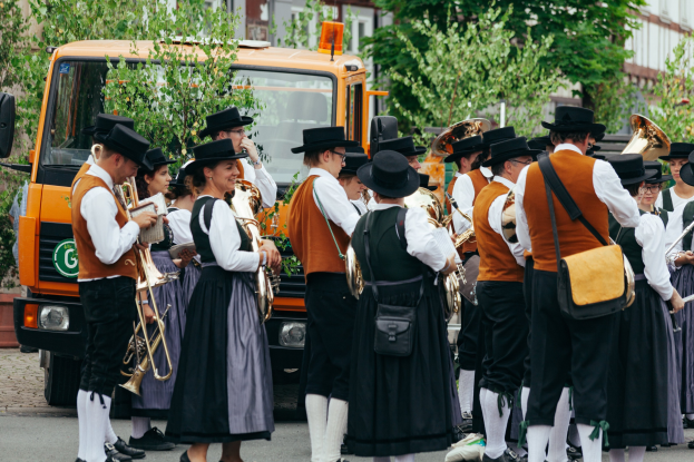 Gruppe von Menschen in traditioneller bayrischer Tracht, die mit Instrumenten marschieren und Taschen tragen, mit Bäumen und Gebäuden im Hintergrund und einem geparkten Fahrzeug am Straßenrand.