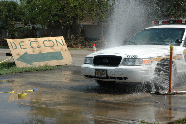 Ein Polizeiwagen fährt durch eine Pfütze, mit einer Tafel, Verkehrskegel und anderen Gegenständen am Boden, Bäume und Gebäude im Hintergrund unter einem klaren blauen Himmel.