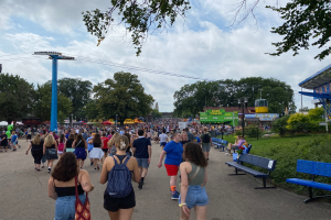 Eine Menschenmenge, die eine von Bänken, Pflanzen, Gras, Bäumen, Pfosten, Lampen, Drähten und einem Schuppen gesäumte Straße entlanggeht, wobei einige Taschen tragen, unter einem bewölkten Himmel.