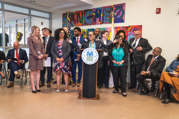 Gruppe von Menschen vor einem Rednerpult mit Mikrofon und einer Tafel mit der Aufschrift "Sancho City Councilman Joins City Council", mit sitzenden und stehenden Teilnehmern, Flaggen, Feuerlöscher und Deckenleuchten im Hintergrund.