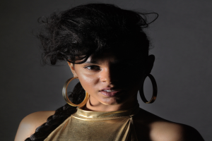 Eine Frau in einem goldenen Kleid und großen Ohrringen steht selbstbewusst vor einer Wand, ihr glattes Bob-Haar und das natürliche Make-up unterstreichen ihren entschlossenen Gesichtsausdruck, während sie direkt in die Kamera blickt.