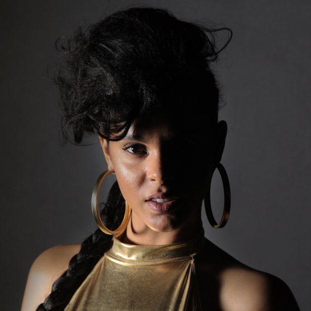 Eine Frau in einem goldenen Kleid und großen Ohrringen steht selbstbewusst vor einer Wand, ihr glattes Bob-Haar und das natürliche Make-up unterstreichen ihren entschlossenen Gesichtsausdruck, während sie direkt in die Kamera blickt.