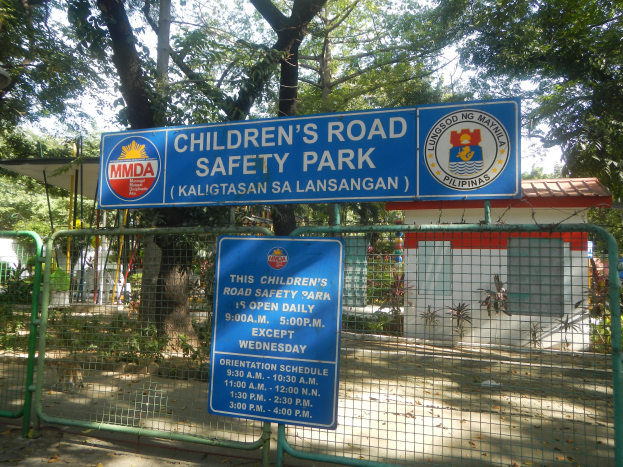 Metallzaun um einen Kinderstraßensicherheitspark mit zwei blauen Informationsschildern, einem kleinen Haus, Pflanzen, Bäumen und einem bewölktem Himmel.