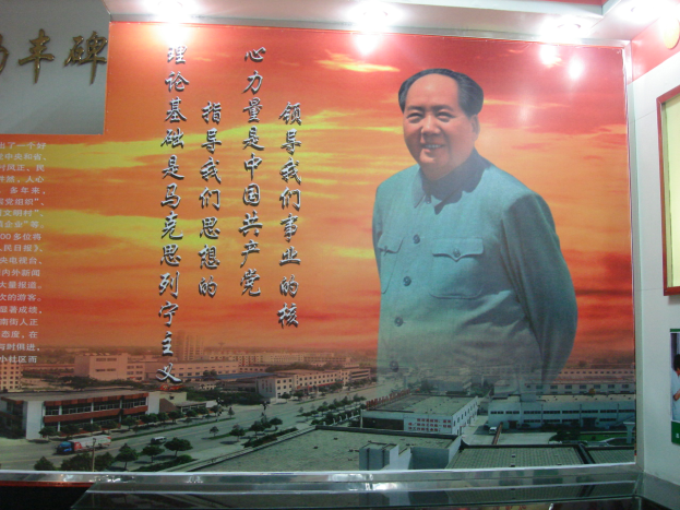 Ein großes beleuchtetes Plakat von Mao Zedong an einer Gebäudewand, mit einem Foto-Rahmen und Text rechts daneben.
