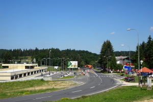 Ein Kreisverkehr mit Fahrzeugen, Gras, Laternen, Texttafeln, Gebäuden, Bäumen und einem bewölkten Himmel, mit einem Straßenschild im Vordergrund.