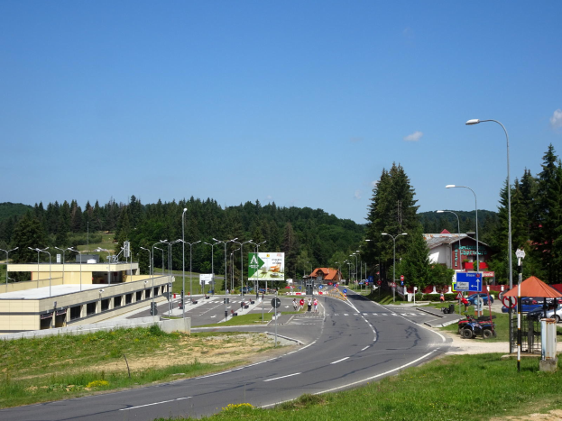 Ein Kreisverkehr mit Fahrzeugen, Gras, Laternen, Texttafeln, Gebäuden, Bäumen und einem bewölkten Himmel, mit einem Straßenschild im Vordergrund.