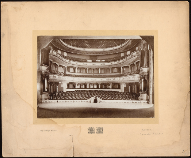 Schwarzes und weißes Foto des Auditoriums der Royal Albert Hall in London, das die Bühne, Reihen von Stühlen, Säulen, Geländer und Text mit einem Logo unten zeigt.