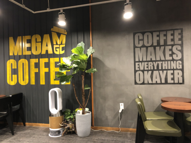 Ein Kaffeehaus-Interieur mit Tischen, Stühlen, einem "Mega Coffee"-Schild an der Wand, einem Topf mit einer Pflanze, einem Bodenständer und Deckenleuchten.