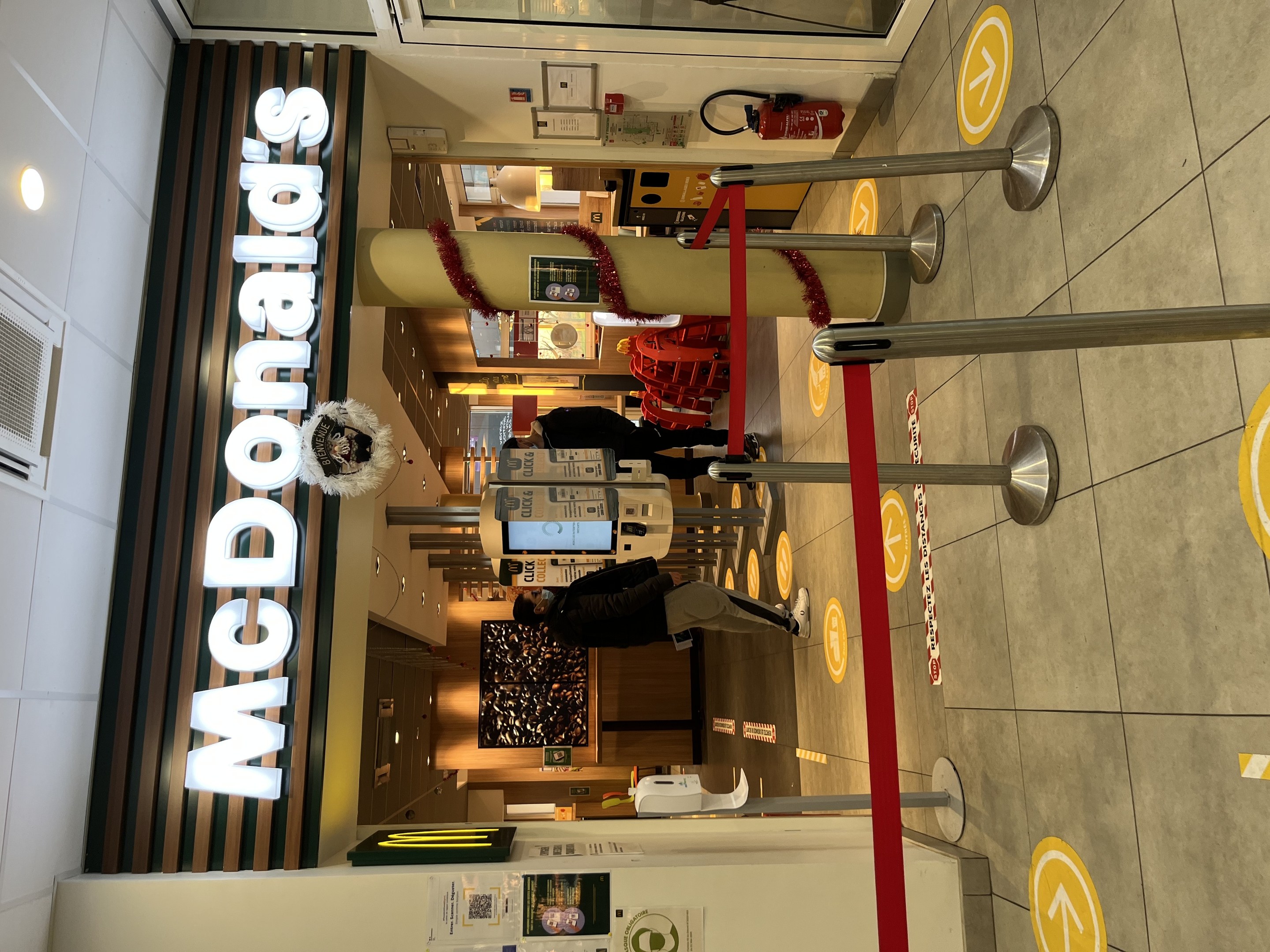 Innenansicht eines McDonald's-Restaurants mit einem sichtbaren Schild, Metallstangen mit roten Bändern auf der rechten Seite und einigen Menschen in der Nähe, wobei im Hintergrund Gegenstände teilweise sichtbar sind.