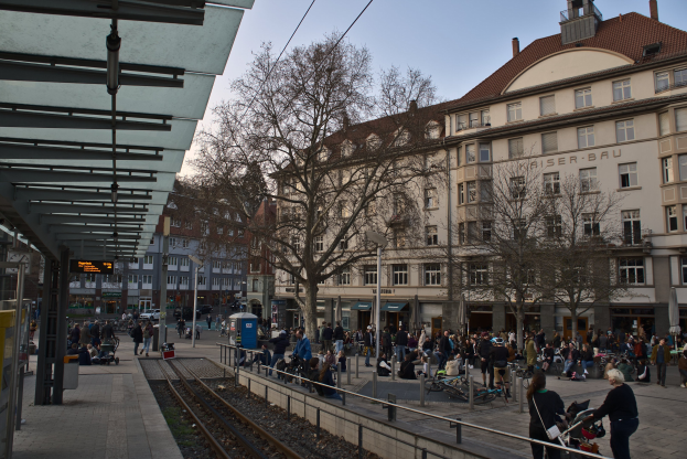 Eine belebte Straßenkreuzung mit einem Bahnhof auf der linken Seite, umgeben von Gebäuden, Bäumen, Laternen und Texttafeln, mit Menschen, die gehen, auf Bänken sitzen und stehen, und dem Himmel im Hintergrund.