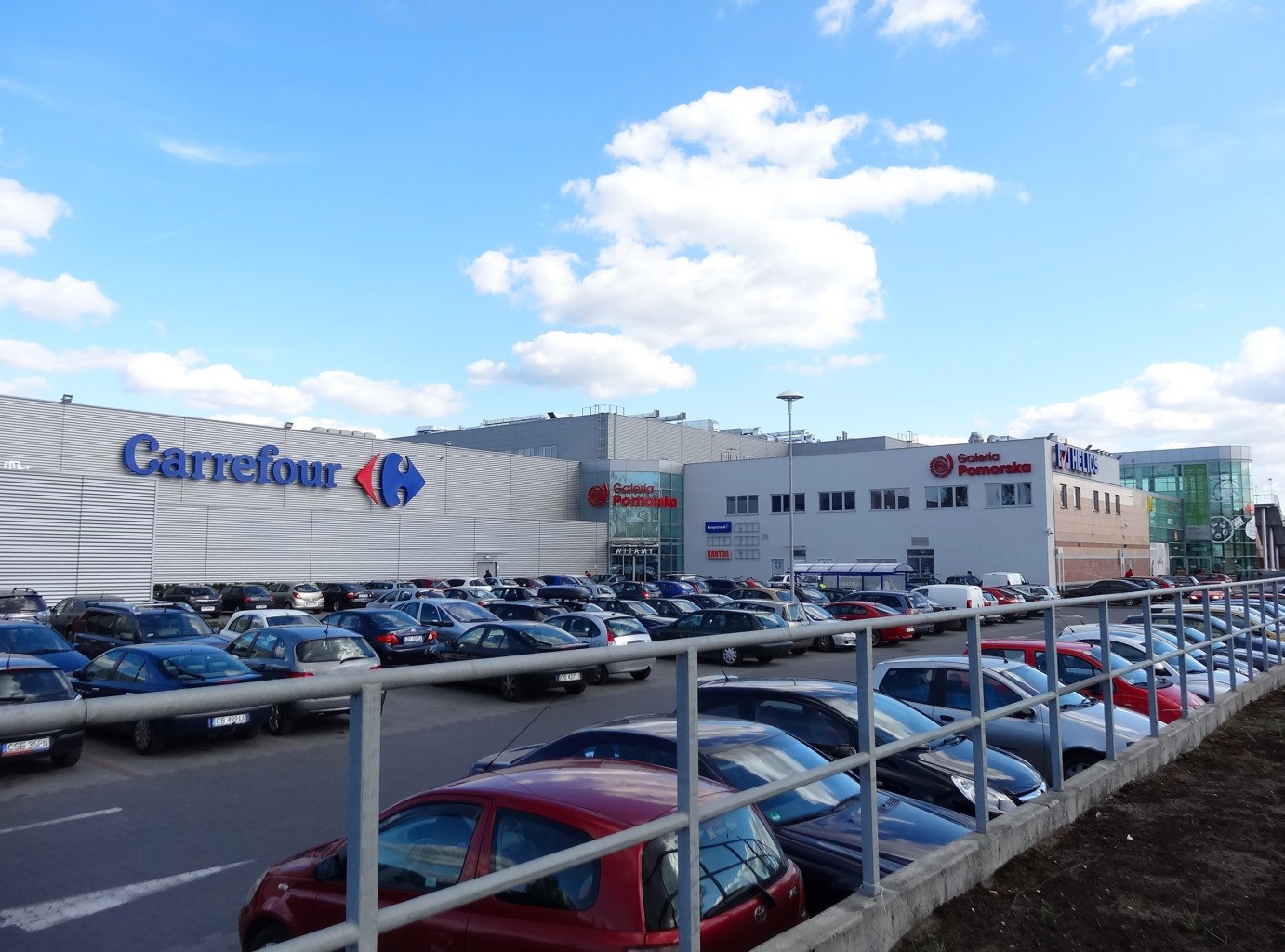 Großer Autohandel mit parkenden Autos vorne, umgeben von Gebäuden, Straßenlaternen, Schildern, fahrenden Fahrzeugen und einem bewölkten Himmel.