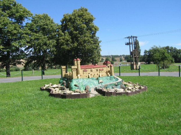 Minigolf-Platz auf einer Rasenfläche, umgeben von einer Kettenzaun-Umzäunung, mit Bäumen, Strommasten und einem bewölkten Himmel im Hintergrund.