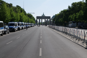 Lange Reihe von Polizeiwagen, die auf der Seite einer Straße vor dem Reichstagsgebäude in Berlin, Deutschland, geparkt sind, mit Menschen auf Fahrrädern und auf der Straße stehend, Barrieren, Bäume, ein Bogen mit Statuen im Hintergrund und sichtbarem Himmel.