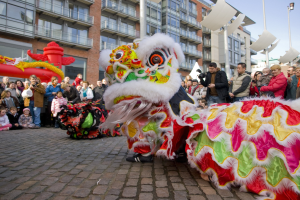 Ein lebendiges chinesisches Neujahrsfest in Amsterdam mit einem Löwen tanzen im Vordergrund und einer Menschenmenge drumherum, einige halten Kameras, vor einem Hintergrund aus Gebäuden, Laternenmasten und einem klaren blauen Himmel.