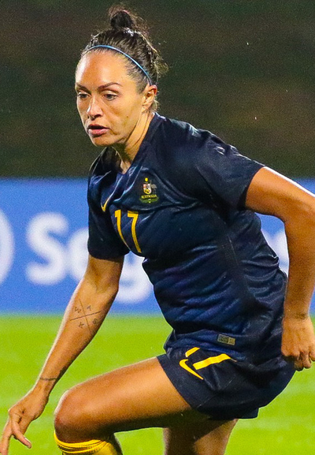 Eine Fußballspielerin in Aktion auf dem Feld, die ein blaues T-Shirt und gelbe Shorts trägt, mit einem unscharfen Hintergrund, der eine blaue Tafel mit Text zeigt.