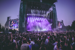 Eine große Menge bei einem Musikfestival schaut Performern auf einer beleuchteten Bühne mit Instrumenten und Bildschirmen zu, vor einem Gebäude, Bäumen und einem klaren Himmel.