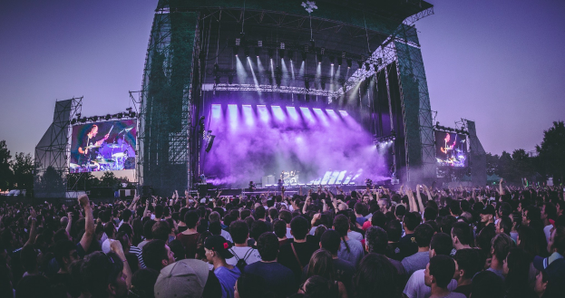 Eine große Menge bei einem Musikfestival schaut Performern auf einer beleuchteten Bühne mit Instrumenten und Bildschirmen zu, vor einem Gebäude, Bäumen und einem klaren Himmel.