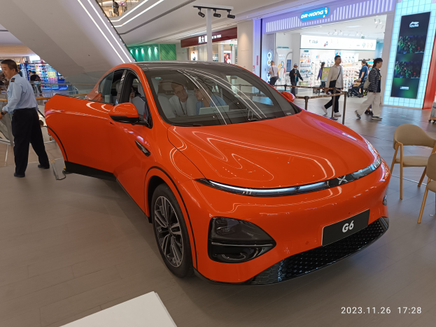 Oranger Elektroauto in einer Ausstellungshalle mit einer Gruppe von Menschen drumherum, Stühle und einen Wagen rechts, Geschäfte und Schilder im Hintergrund.