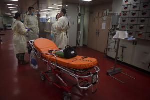 Gruppe von Menschen in Schutzausrüstung um ein Bett in einem Krankenhauszimmer mit medizinischer Ausrüstung, einer Trage, einem Ständer, einer Tür, einer Säule, einem Schild und Deckenleuchten.