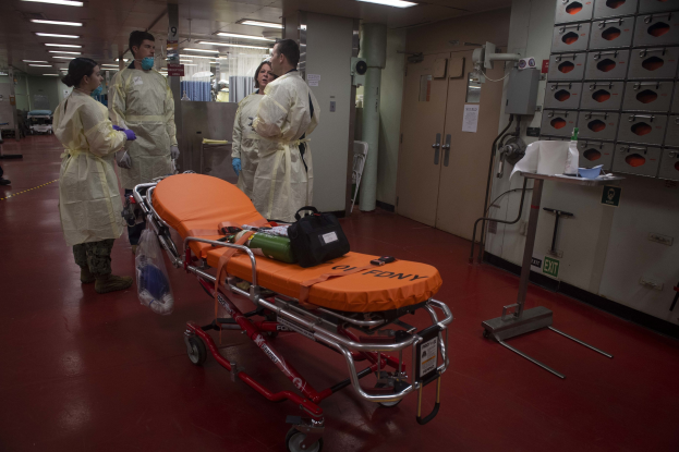 Gruppe von Menschen in Schutzausrüstung um ein Bett in einem Krankenhauszimmer mit medizinischer Ausrüstung, einer Trage, einem Ständer, einer Tür, einer Säule, einem Schild und Deckenleuchten.