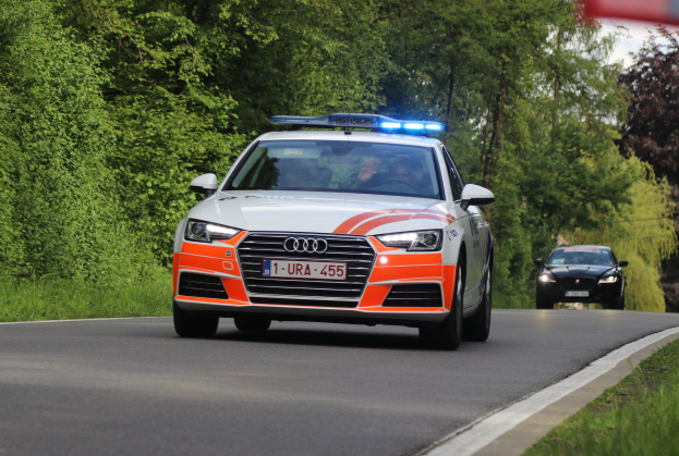 Polizeifahrzeug (Audi A4) fährt auf einer grasbewachsenen Straße mit zwei Insassen, umgeben von Bäumen unter einem klaren blauen Himmel.