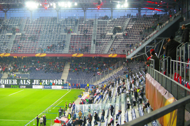 Großes Stadion voller Zuschauer bei einem Fußballspiel, mit sitzenden und stehenden Personen, einem Geländer, einem Banner auf der rechten Seite und einem leeren Abschnitt im Hintergrund mit Deckenbeleuchtung.