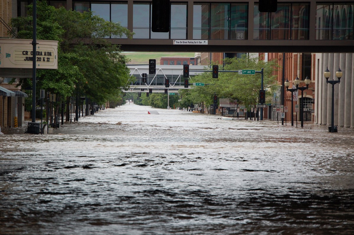 Überflutete Stadtstraße mit Wasser bedeckter Straße, Infrastruktur und Gebäuden sowie eine Brücke im Hintergrund.