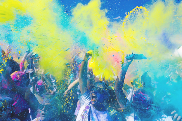 Menschen feiern Holi, werfen farbiges Pulver gegen einen hellblauen Himmel.